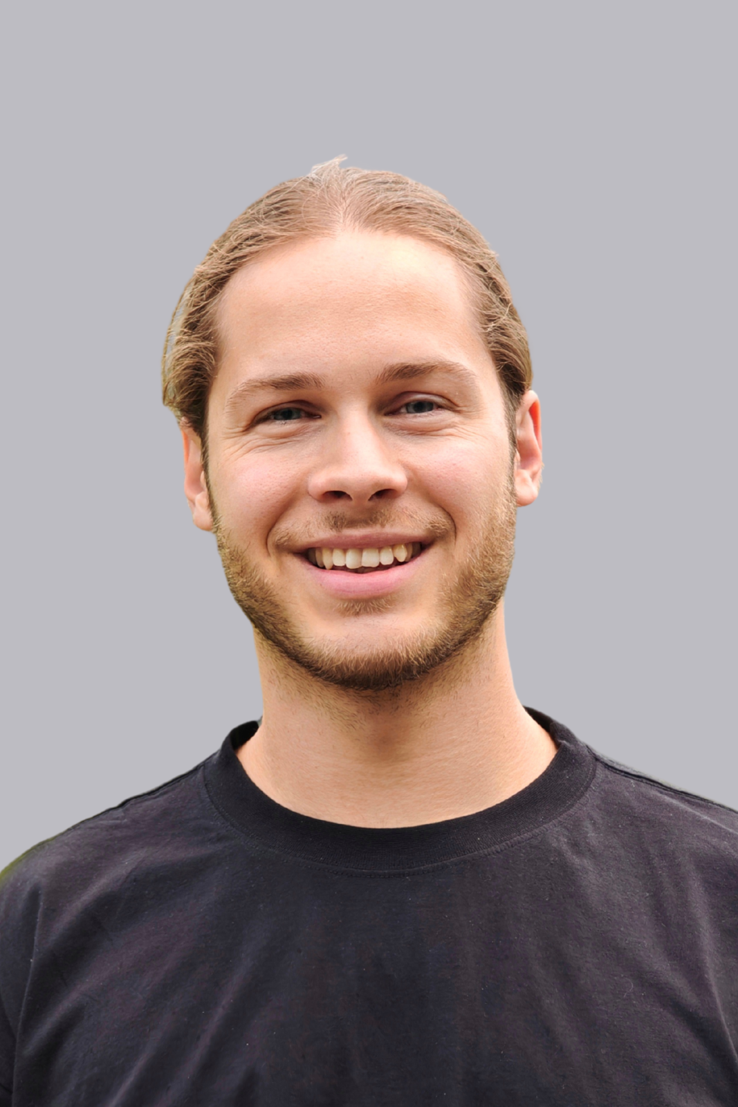 Un jeune homme aux cheveux châtain clair, portant une barbe et un t-shirt noir, sourit devant un fond gris.