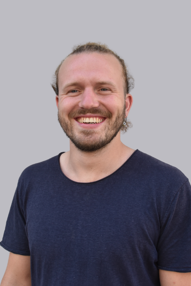 Un homme souriant aux cheveux longs, vêtu d'un t-shirt bleu, sur fond gris.