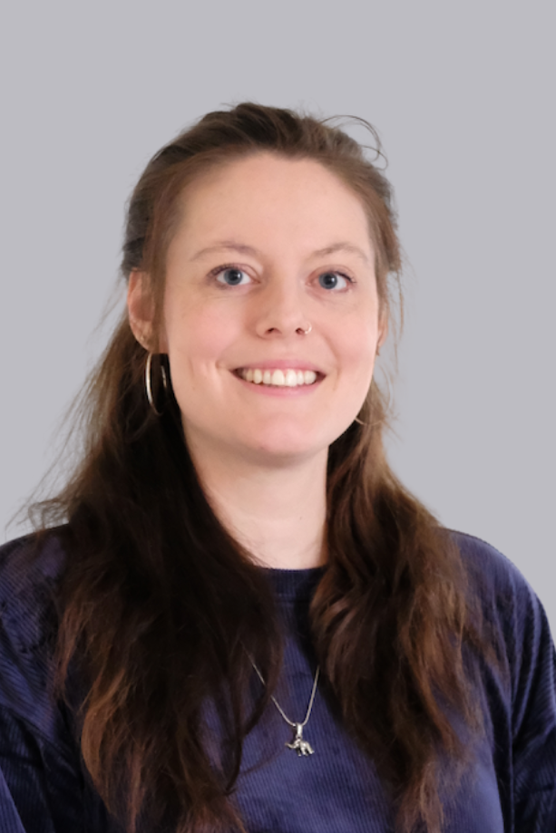 Jeune femme aux longs cheveux bruns, avec un piercing au nez et une boucle d'oreille, vêtue d'un pull bleu, souriante devant un fond neutre.